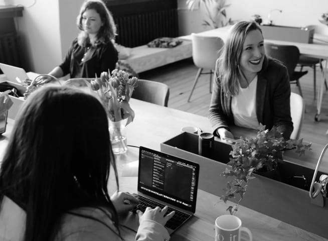 Three women are diligently working: one is engrossed in her phone, chuckling at something to her right, while the other is focused on her laptop.